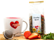 Eine Tasse mit einem roten Herz und der Aufschrift „Verliebt in Halle“ steht neben einem Tee-Ei, frischen Erdbeeren, Minze und einem durchsichtigen Beutel Tee „Frischer Wind“ mit Blumenmotiv – alles auf einer Holzoberfläche vor einem weißen Hintergrund.