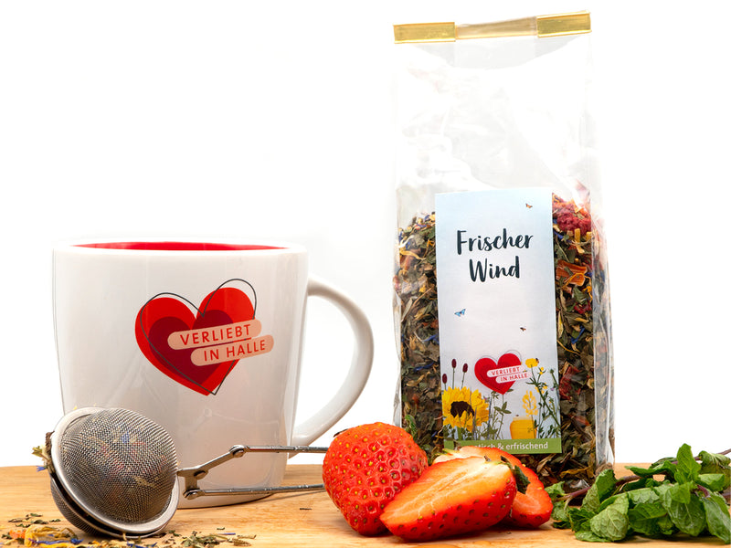 Eine Tasse mit einem roten Herz und der Aufschrift „Verliebt in Halle“ steht neben einem Tee-Ei, frischen Erdbeeren, Minze und einem durchsichtigen Beutel Tee „Frischer Wind“ mit Blumenmotiv – alles auf einer Holzoberfläche vor einem weißen Hintergrund.