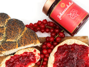 Eine Mohnbrotscheibe und eine runde Brotscheibe, beide mit dem leuchtenden Fruchtaufstrich „Johannisbeerliebe“ Johannashaller belegt, sind mit frischen roten Johannisbeeren garniert. Das Glas steht auf einem Holzbrett vor weißem Hintergrund.
