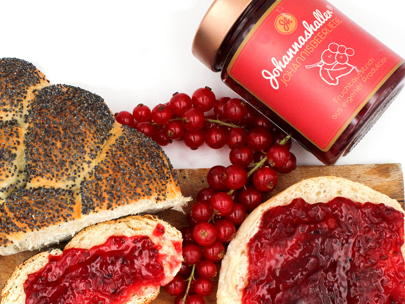Eine Mohnbrotscheibe und eine runde Brotscheibe, beide mit dem leuchtenden Fruchtaufstrich „Johannisbeerliebe“ Johannashaller belegt, sind mit frischen roten Johannisbeeren garniert. Das Glas steht auf einem Holzbrett vor weißem Hintergrund.