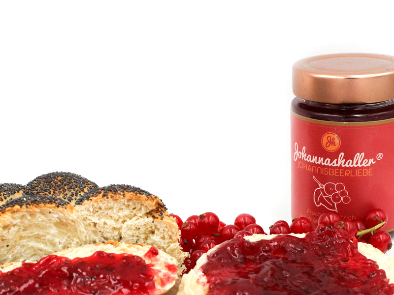Rechts steht ein Glas Fruchtaufstrich „Johannisbeerliebe“ Johannashaller, neben Mohnbrot und Marmeladenbrötchen. Vorne sind frische rote Johannisbeeren verstreut, die vor weißem Hintergrund die Regionalität betonen.