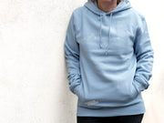 Eine Person steht vor einer weißen strukturierten Wand und trägt den „Verliebt in Halle“-Skyline-Halle-Hoodie mit einer minimalistischen Strichzeichnung auf der Brust. Ihre Hände stecken in den Taschen; schwarze Hosen und ein Teil ihres Gesichts sind teilweise sichtbar.