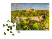 Ein teilweise fertiggestelltes Puzzle „Die Burg Giebichenstein in Halle (Saale)“ zeigt eine Steinbrücke über einen Fluss, grüne Bäume und eine historische Festung auf einem Hügel, wobei mehrere lose Teile auf einem weißen Hintergrund verstreut sind.